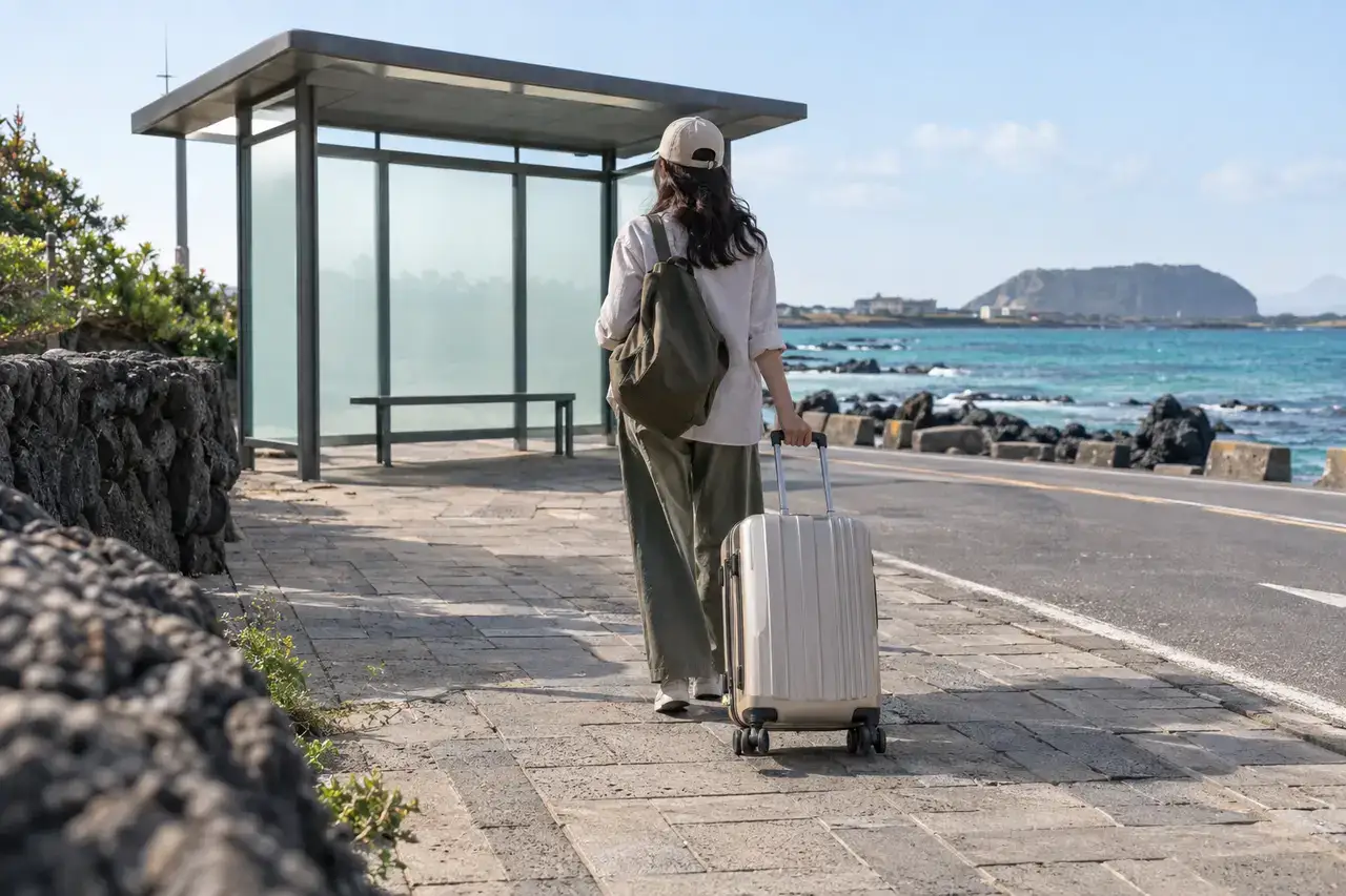 Traveler rolling a suitcase toward a blank coastal bus shelter beside turquoise water and basalt walls in northeast Jeju.