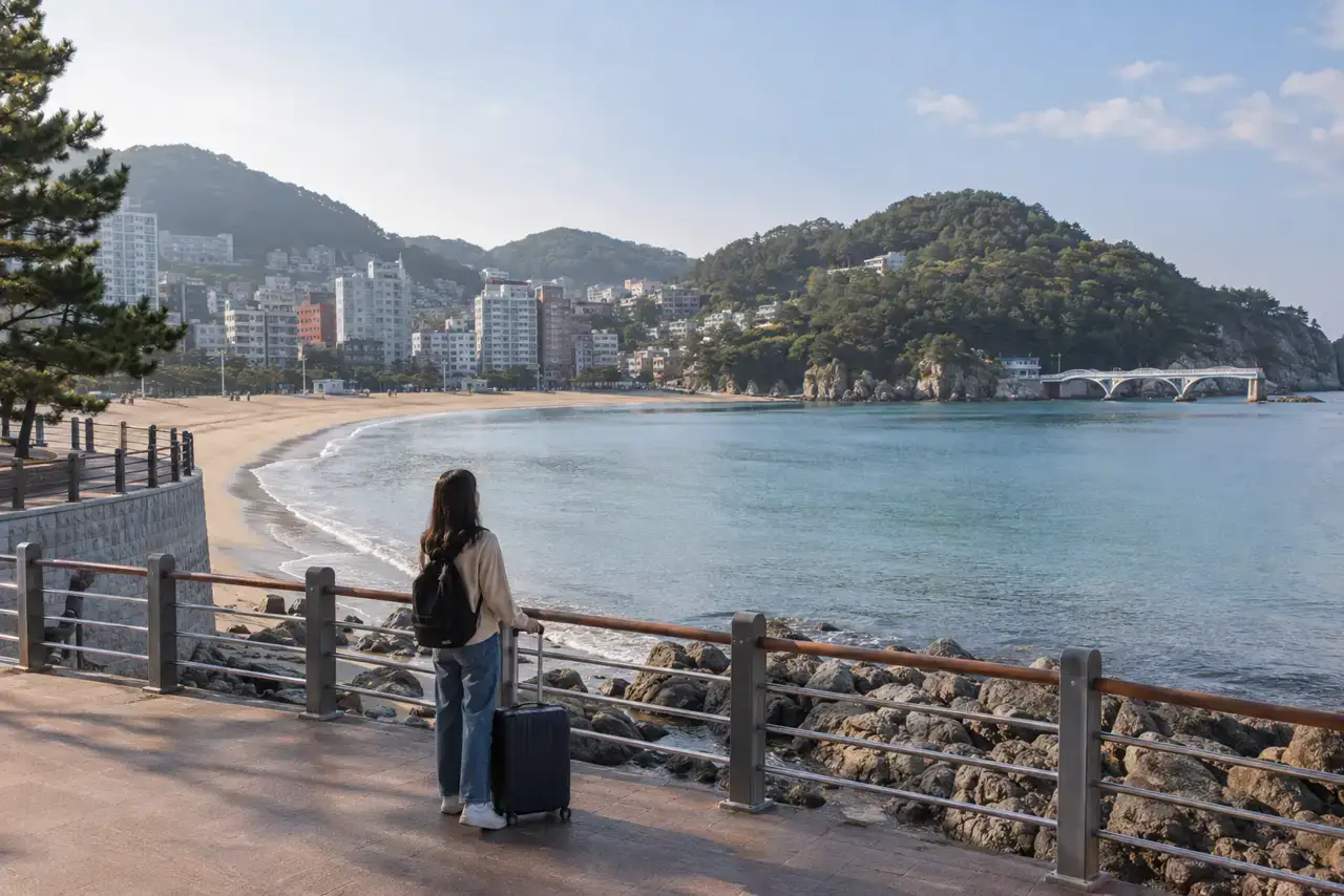 A traveler with a suitcase on the public promenade above Songdo Beach in Busan on a quiet autumn morning.