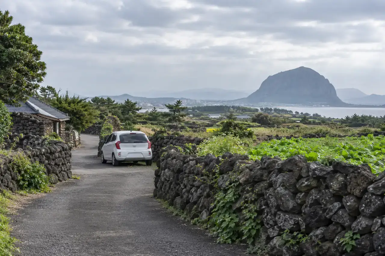 A compact car on a narrow basalt-wall lane in Andeok with Sanbangsan in the distance.