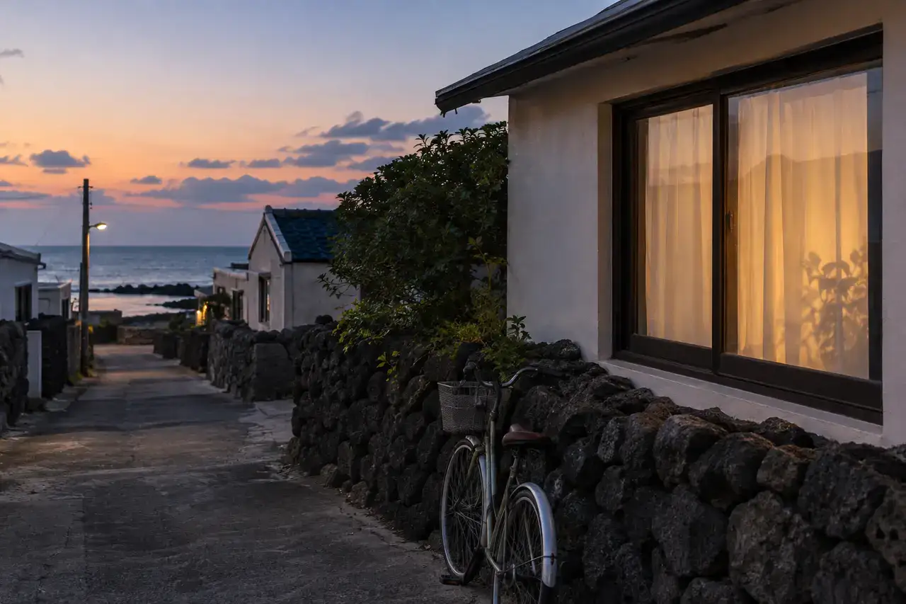 Quiet Sehwa village lane at blue hour with basalt walls, a bicycle and a softly lit curtained window near the sea.