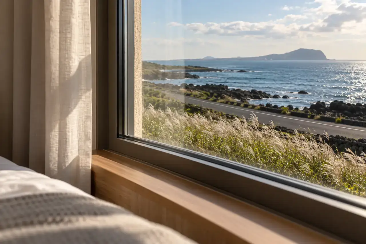 Closed guest-room window overlooking a Jeju coastal road, silver grass and the sea.